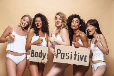 İç çamaşırlı, mutlu, çok uluslu kadınların bej arka plan üzerinde başparmaklarını gösteren ve pankartlarını tutan resimleri.