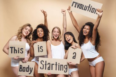 Heyecanlı, iç çamaşırlı, gülümseyen ve bej arka planda izole edilmiş pankartları tutan çok uluslu kadınların görüntüsü