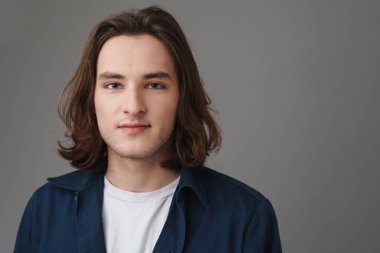 Gri arka planda izole edilmiş, uzun saçlı, poz veren ve kameraya bakan çekici bir genç resmi.