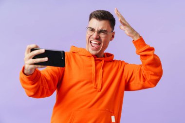 Mor arka planda yalnız poz veren mutlu genç adamın fotoğrafı cep telefonuyla selfie çekin..