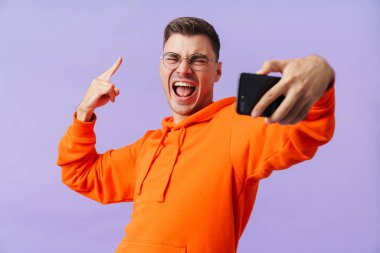 Mor arka planda yalnız poz veren mutlu genç adamın fotoğrafı. Kaya hareketini gösteren cep telefonuyla bir selfie çek..