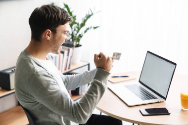 Evde dizüstü bilgisayarla ödeme yaparken gülümseyen ve elinde kredi kartı tutan mutlu bir genç resmi