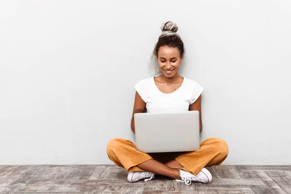 穿着休闲装 孤零零地坐在白色背景上 膝上抱着手提电脑的年轻非洲女人笑着 — 图库照片