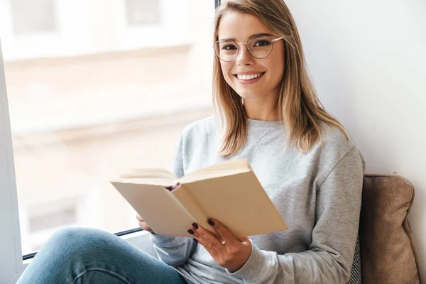 Gözlüklü Gülümseyen Güzel Bir Kadının Pencerenin Yanında Otururken Kitap Okuduğu — Stok fotoğraf