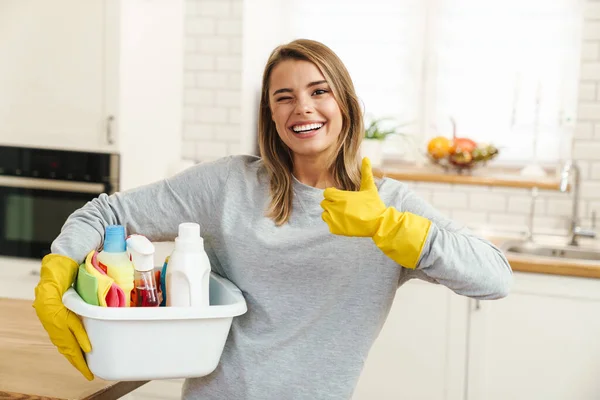 Photo of smiling young woman housewife in gloves holding cleanser bottles and showing thumb up at modern kitchen