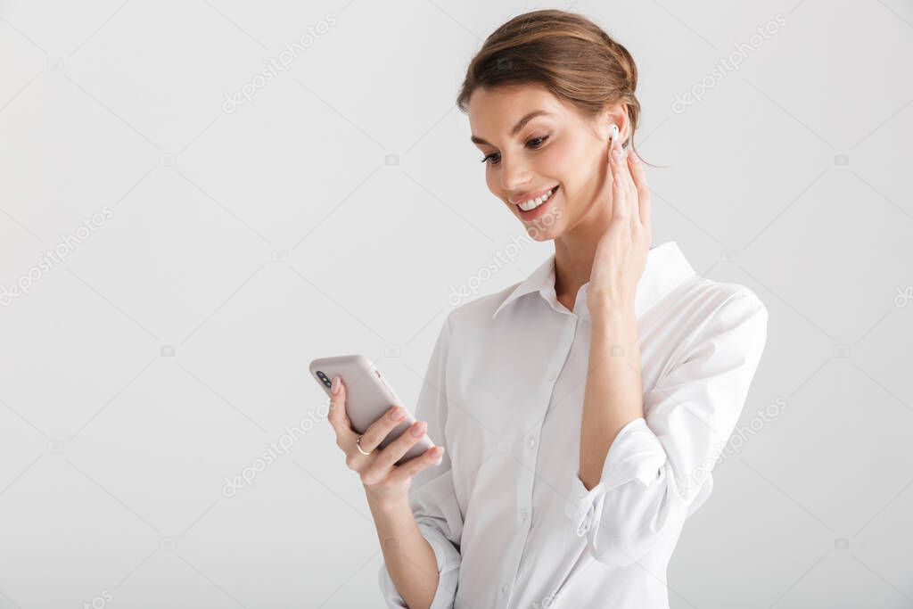 Image of smiling beautiful woman using wireless earphone and mobile phone isolated over white background
