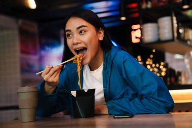 Neşeli bir Asyalı kadının Çin eriştesi yerken ve kafede otururken gülümsediği fotoğraf.