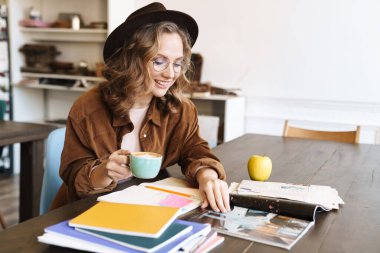 Gözlüklü neşeli bir kadının evde egzersiz kitaplarıyla çalışırken kahve içtiği görüntüsü.