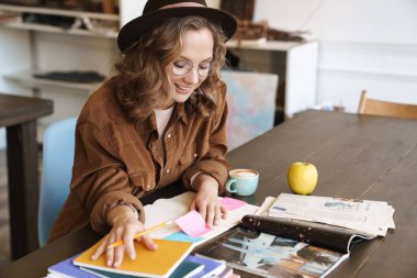 Gözlüklü ve şapkalı neşeli çekici bir kadının evde alıştırma kitaplarıyla çalışırken gülümsemesi.