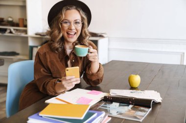 Gözlüklü ve şapkalı gülümseyen bir kadının evde kahve içerken cep telefonu kullanması.