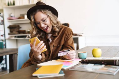 Evde ders çalışırken cep telefonu ve kredi kartı kullanan şapkalı heyecanlı bir kadın resmi.