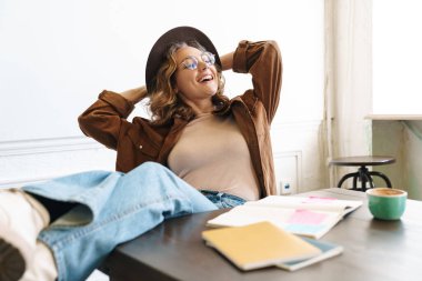 Evde ders çalışırken bacakları masada dinlenen şapkalı genç bir kadın resmi.