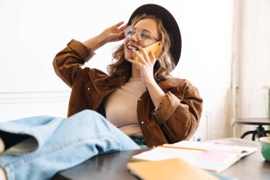 Evde ders çalışırken cep telefonuyla konuşan şapkalı, gülümseyen genç bir kadın.