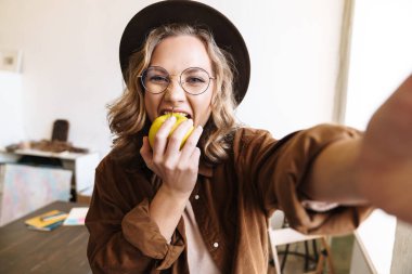Şapkalı, gülümseyen ve evde fotoğraf çekerken elma tutan neşeli genç bir kadın resmi.
