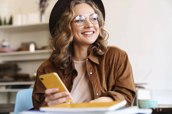 Obrázek Usměvavé Ženy Brýlích Klobouku Pomocí Mobilního Telefonu Při Studiu — Stock fotografie