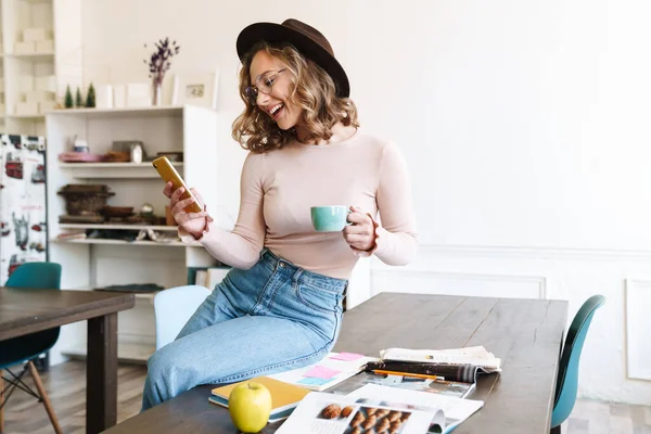 自宅のテーブルに座りながら携帯電話やコーヒーを飲んでいる陽気な若い女性のイメージ — ストック写真