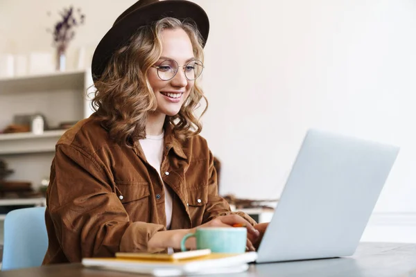 テーブルに座りながらノートパソコンで働く眼鏡と帽子の明るい女性のイメージ — ストック写真