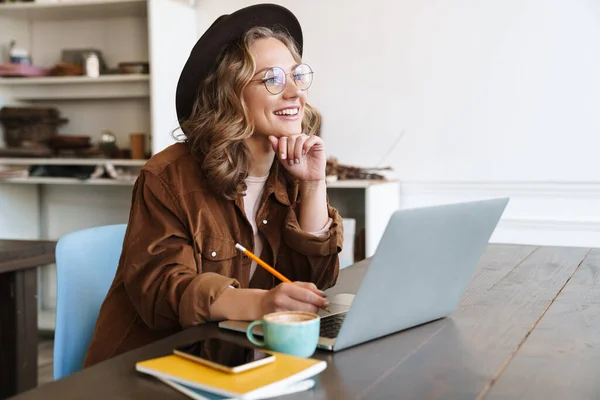 Gözlüklü Şapkalı Neşeli Bir Kadının Masada Otururken Dizüstü Bilgisayarla Çalışması — Stok fotoğraf