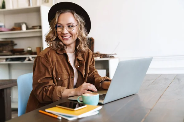 Gözlüklü Şapkalı Neşeli Bir Kadının Masada Otururken Dizüstü Bilgisayarla Çalışması — Stok fotoğraf