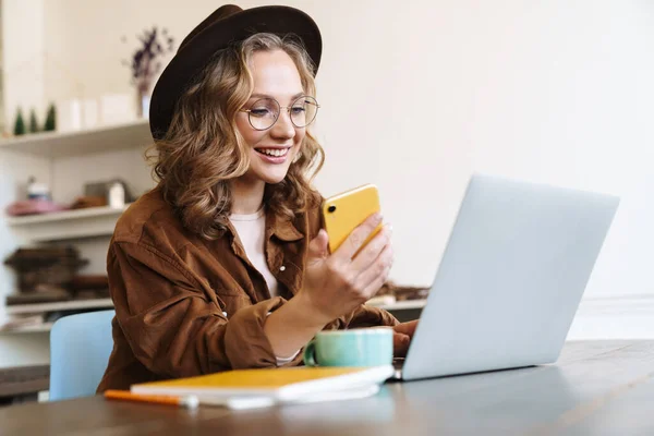 テーブルに座っている間 ノートパソコンや携帯電話で働く帽子の明るい若い女性のイメージ — ストック写真