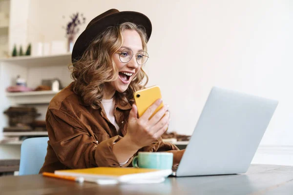 Şapkalı Laptoplu Cep Telefonlu Heyecanlı Genç Bir Kadının Masada Otururken — Stok fotoğraf