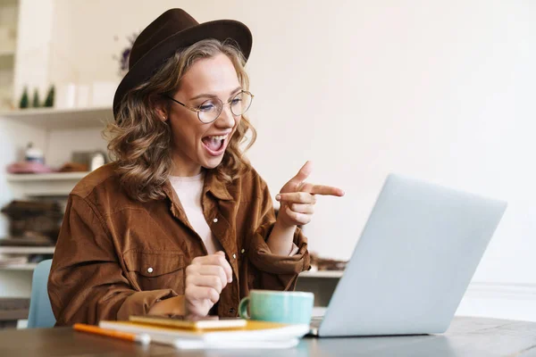 興奮した若い女性のイメージで帽子作業ノートパソコンと指差し指で画面に座ってテーブル — ストック写真