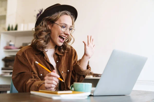 興奮した若い女性のイメージで帽子作業でラップトップと手を振ってテーブルに座って — ストック写真