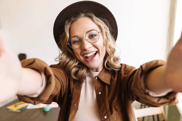 自撮りしながら笑う帽子や眼鏡の中の楽しい若い女性のイメージ室内 — ストック写真