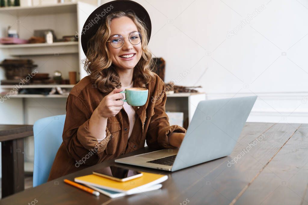 Image of cheerful charming woman in hat working with laptop and drinking coffee while sitting at table