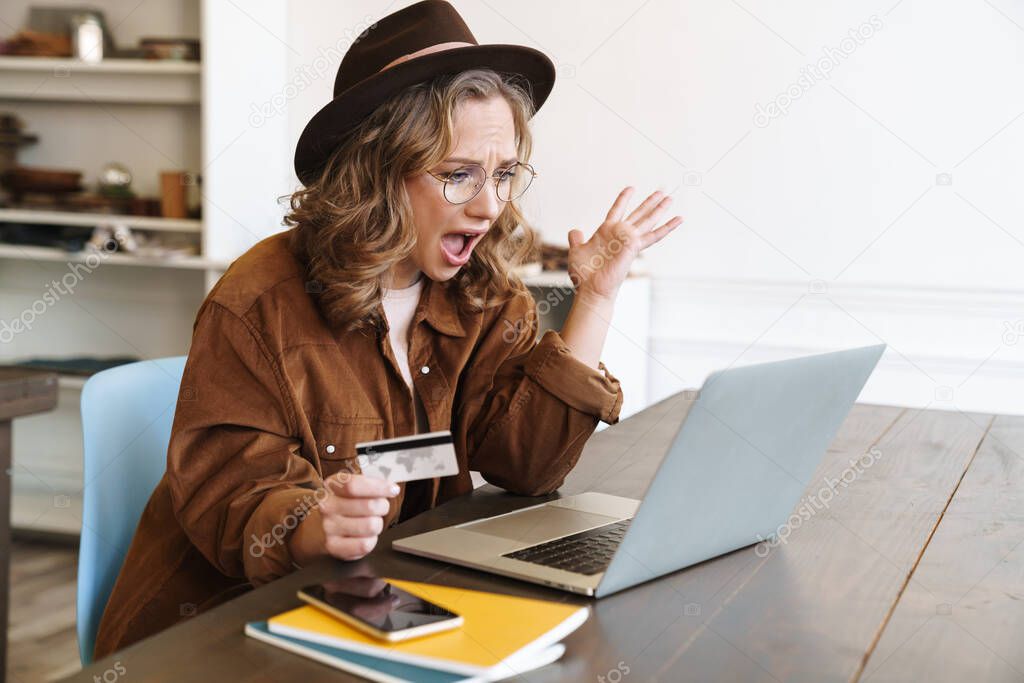Image of angry young woman in hat expressing outrage and holding credit card while working with laptop