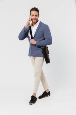 Beyaz arka planda tek başına yürürken cep telefonuyla konuşan ceketli gülümseyen bir genç resmi