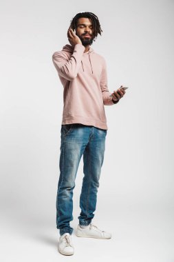 Beyaz arka planda izole edilmiş cep telefonu ve kablosuz kulaklık kullanan memnun Afrikalı Amerikalı adamın portresi.