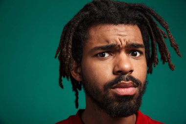 Kafası karışmış, yeşil arka planda tek başına duran ve kameraya bakan, kazak giyen Afrikalı genç bir adamın portresini yakından çek.