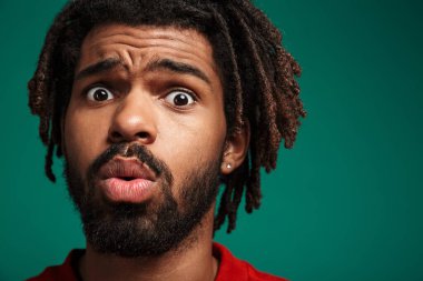 Şok olmuş, yeşil arka planda tek başına duran ve kameraya bakan, kazak giyen Afrikalı genç bir adamın portresini yakından çek.