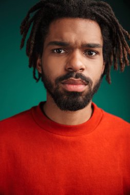 Yeşil arka planda tek başına duran ve kameraya bakan, kazak giyen Afrikalı genç bir adamın yakın portresi.