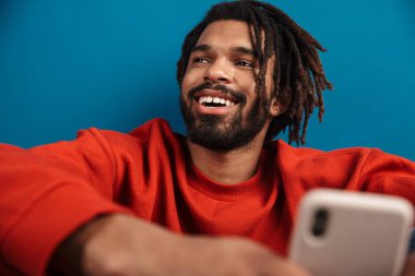 Mavi arka planda tek başına oturan gülümseyen Afrikalı genç bir adamın portresi. Cep telefonu kullanıyor.