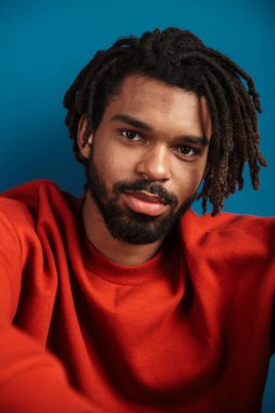 Yeşil arka planda tek başına oturmuş kameraya bakan, kazak giyen Afrikalı genç bir adamın yakın portresi.