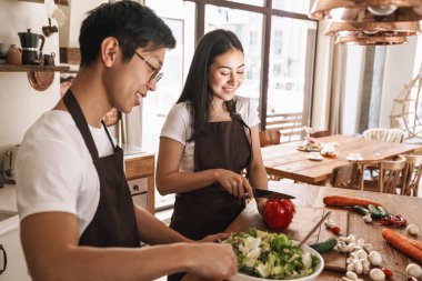 Önlüklü genç, çok kültürlü bir çiftin hoş bir mutfakta gülüp öğle yemeği hazırlayışının resmi.