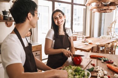 Önlüklü genç, çok kültürlü bir çiftin hoş bir mutfakta gülüp öğle yemeği hazırlayışının resmi.