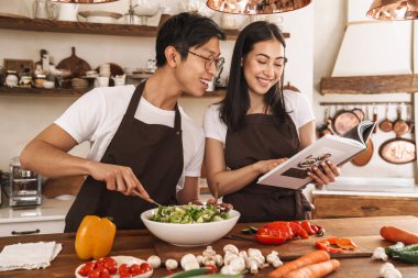 Yemek pişirirken yemek tarifleriyle gülümseyen ve kitap okuyan çok kültürlü bir çiftin görüntüsü.