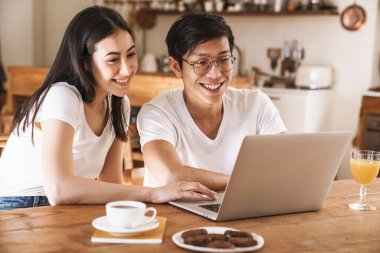 Mutfakta rahat bir masada otururken gülümseyen ve laptop kullanan çok kültürlü mutlu çiftin görüntüsü