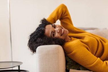 Oturma odasındaki kanepede uzanmış gülümseyen Afro-Amerikan kadını.