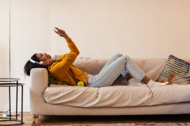 Oturma odasındaki kanepede uzanırken kulaklık ve cep telefonu kullanan neşeli Afro-Amerikan kadın görüntüsü