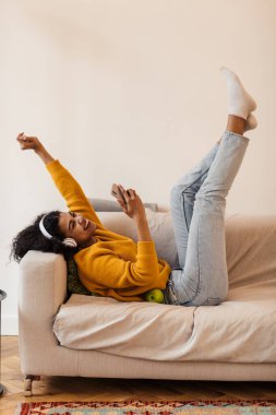 Oturma odasındaki kanepede uzanırken kulaklık ve cep telefonu kullanan neşeli Afro-Amerikan kadın görüntüsü