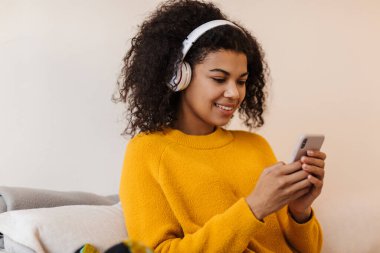 Oturma odasında otururken kulaklık ve cep telefonu kullanan neşeli Afrikalı Amerikalı kadın görüntüsü