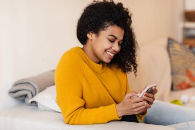 Neşeli bir Afrikalı Amerikalı kadının oturma odasında otururken cep telefonu kullanması ve gülümsemesi.