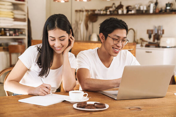 Image of smiling multicultural couple making notes in planner and using laptop in cozy kitchen