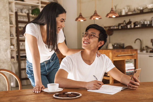 Image of multicultural smiling couple using cellphone and making notes in planner in cozy kitchen