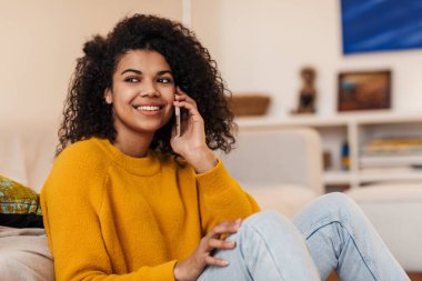 Gülümseyen Afrikalı Amerikalı bir kadının oturma odasında otururken cep telefonuyla konuşması.