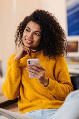 Oturma odasında koltukta otururken cep telefonu kullanan gülümseyen Afrikalı Amerikalı kadın.